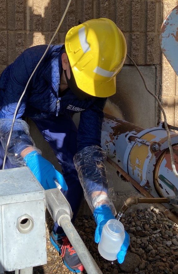 A U.S. Geological Survey technician in a hard hat and gloves filling a water sample bottle for PFAS testing